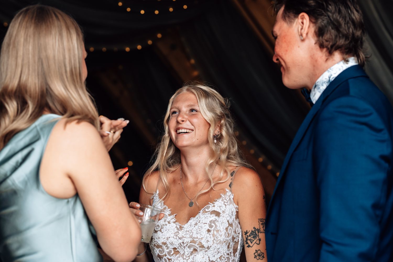 Bride in lace wedding dress laughing with guests at a rustic wedding reception under string lights.