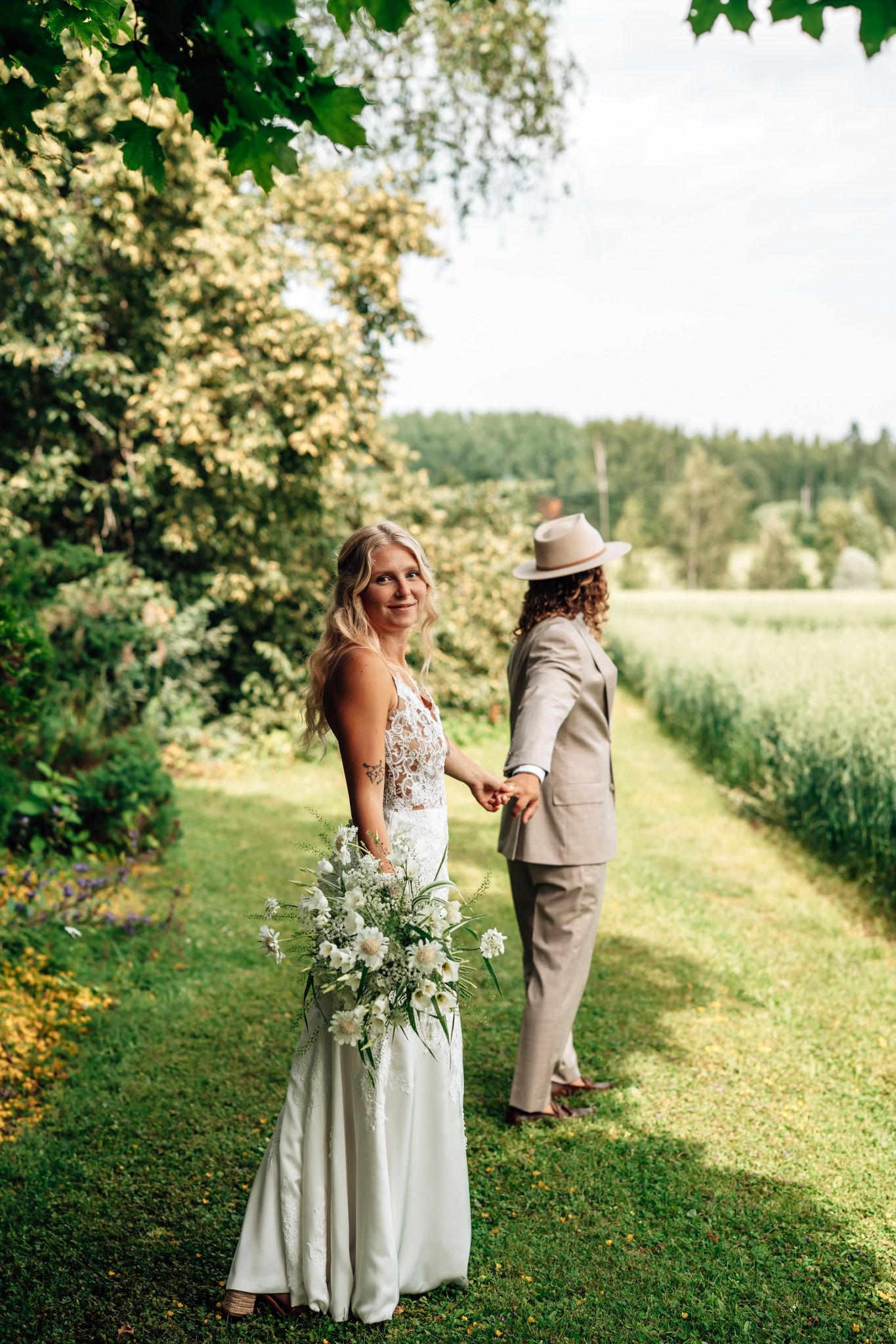 Bride in white gown holding bouquet smiles back while groom in tan suit and hat leads her along lush green garden path.