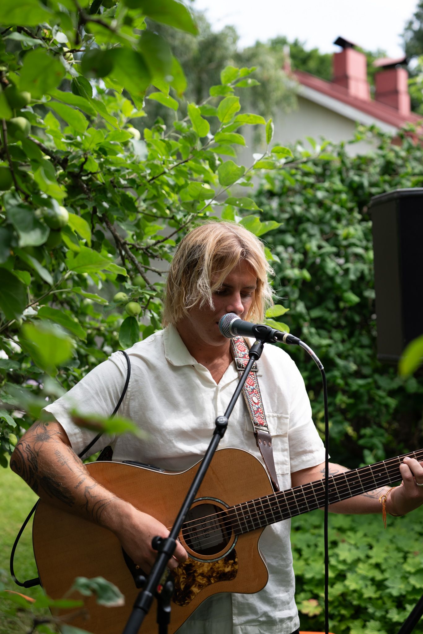 Blonde musician playing acoustic guitar and singing into microphone outdoors in lush green garden setting.