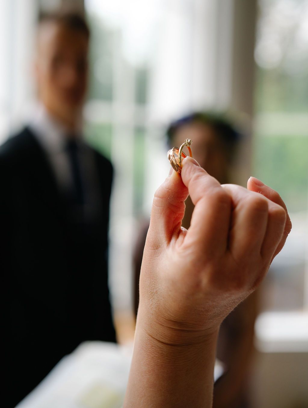 Hand holding two gold wedding rings with blurred groom in background at wedding ceremony.