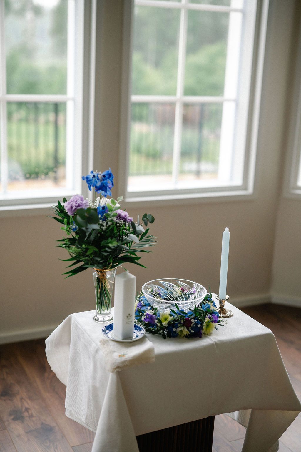 A elegant wedding unity table with white candles, blue floral arrangement, and decorative bowl set near a bright window.