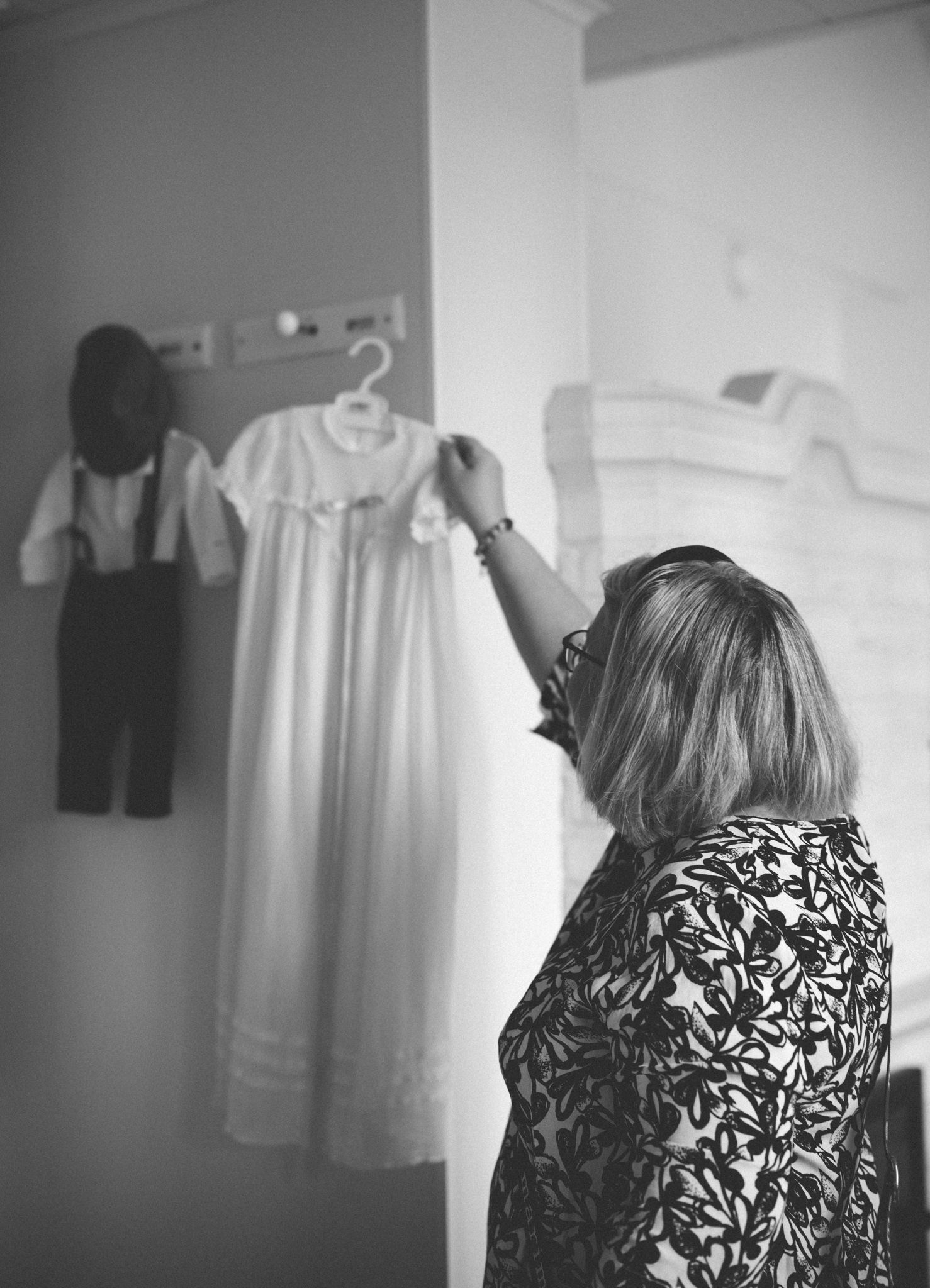 Woman in floral dress hanging white christening gown on wall hook next to boys suit in black and white photo.