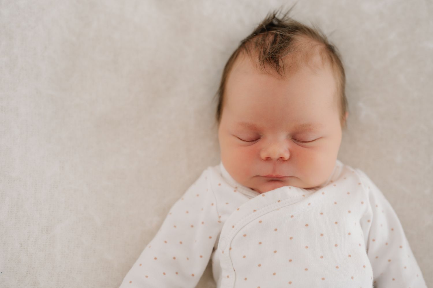 Peaceful newborn baby sleeping on a soft white blanket, wearing a white polka dot onesie with dark hair.