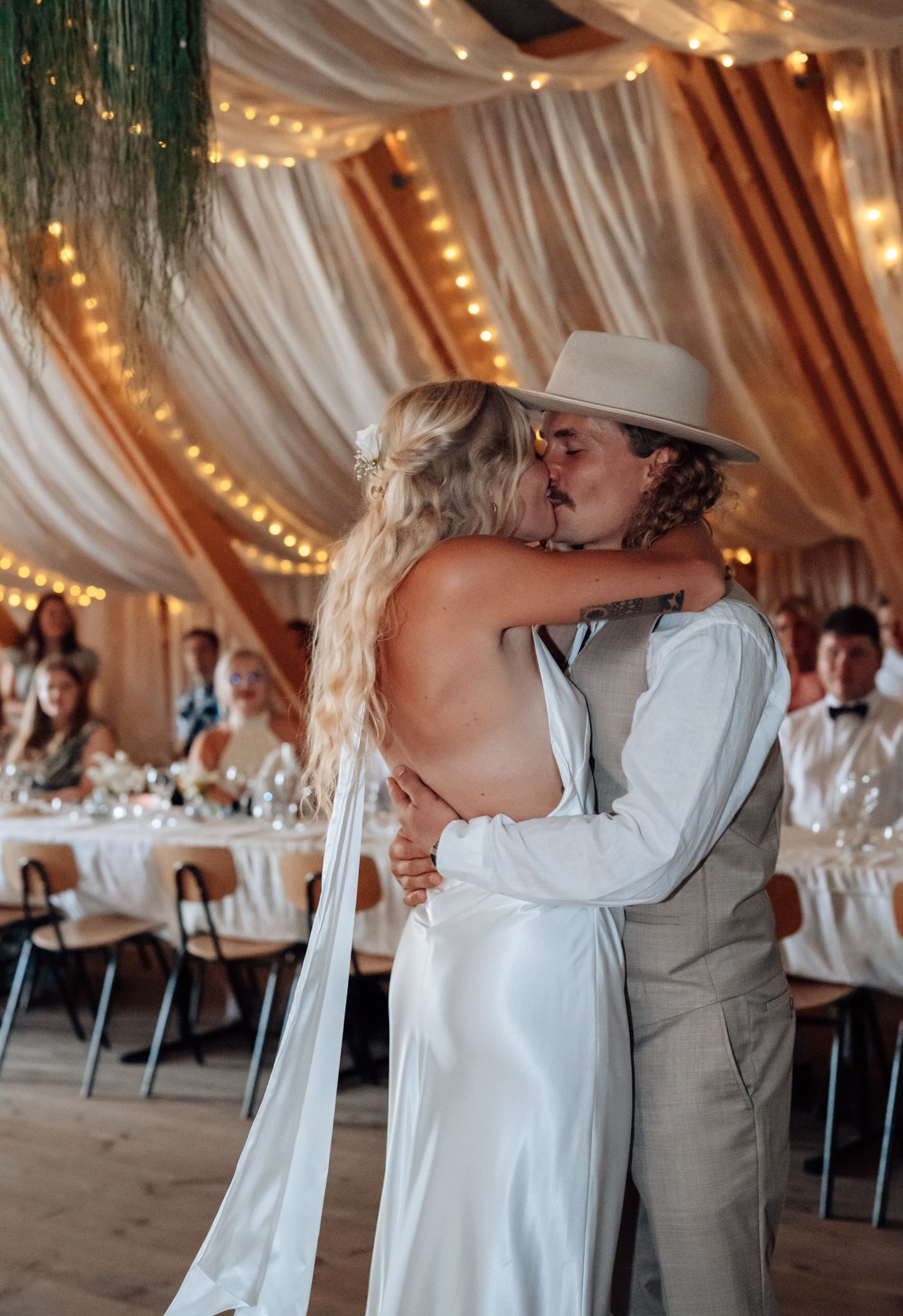 Bride and groom share first kiss at rustic wedding reception with string lights and draped fabric ceiling.