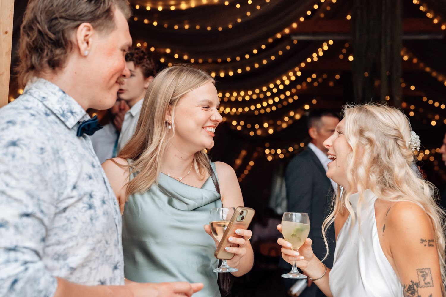 Friends laughing and toasting with champagne glasses at a festive wedding reception with string lights.