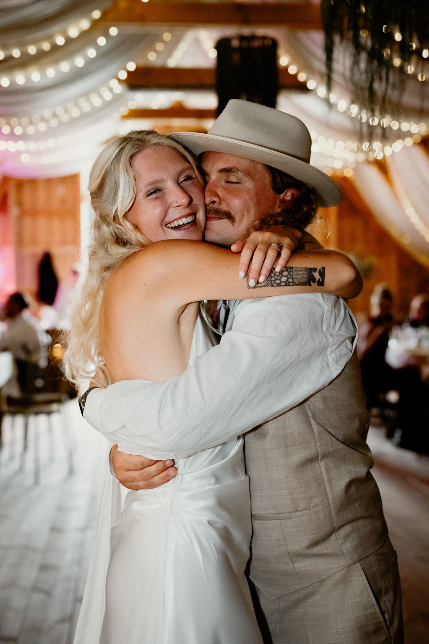 A joyful bride in a white gown hugs a groom in a gray suit and cowboy hat at a warmly lit wedding reception.
