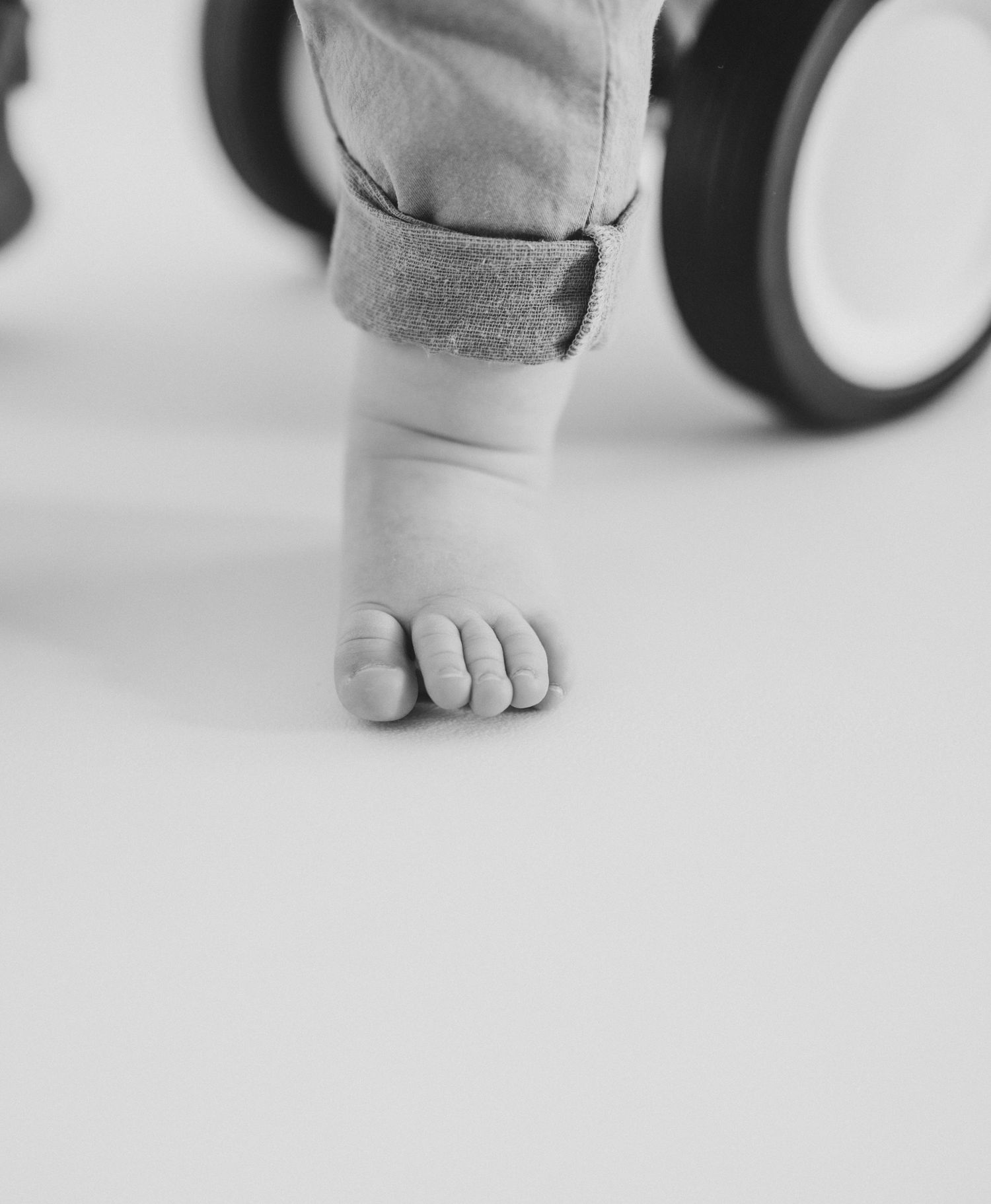 Close-up black and white photo of a baby's bare foot and rolled jeans cuff standing near a stroller wheel.