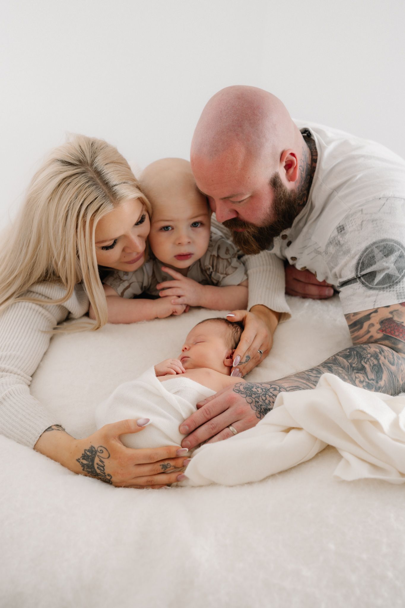 A family of four with tattooed parents and a toddler tenderly gazing at their swaddled newborn on a white fluffy surface.