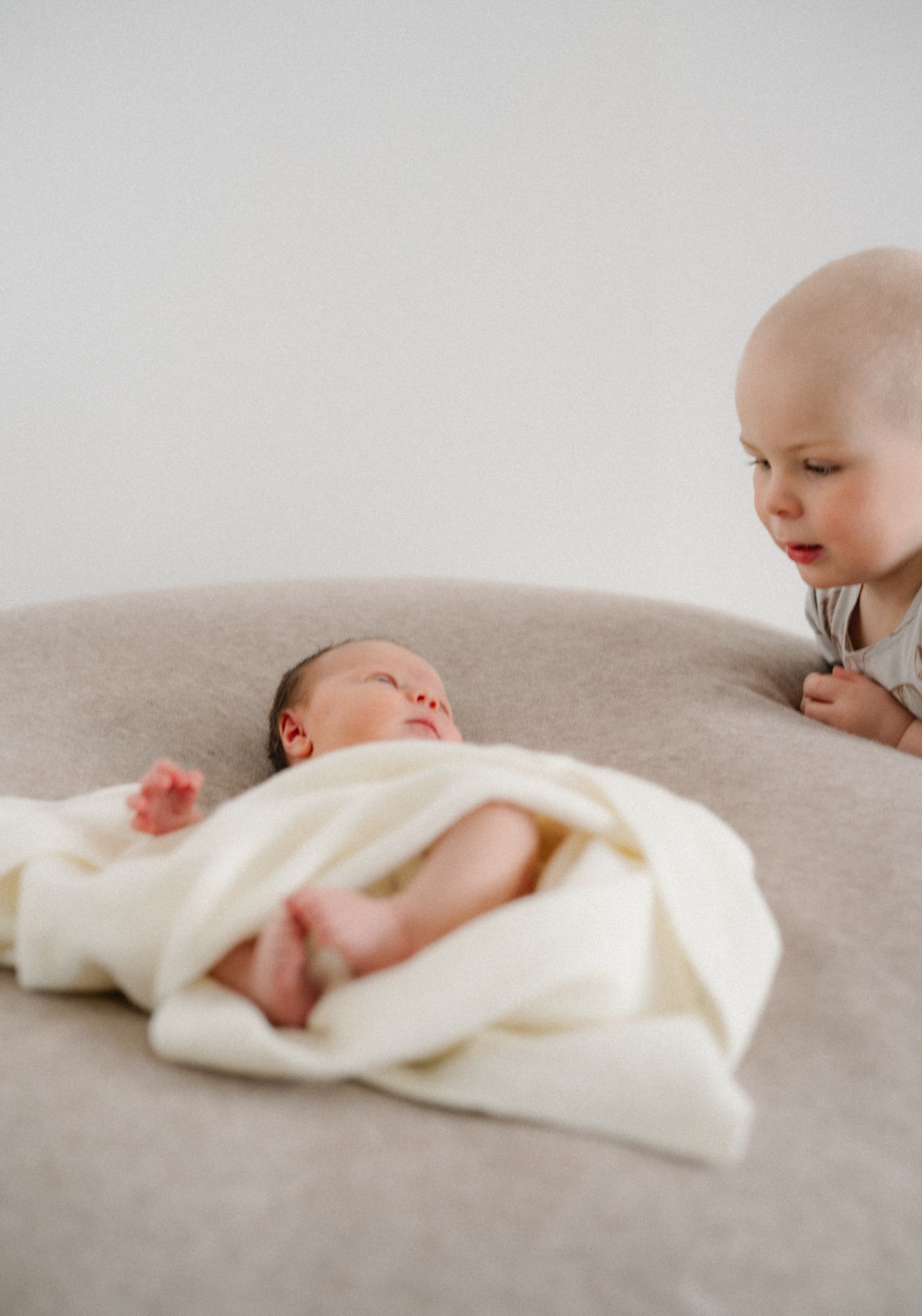 Toddler curiously gazing at sleeping newborn baby wrapped in white blanket on a soft gray cushion.