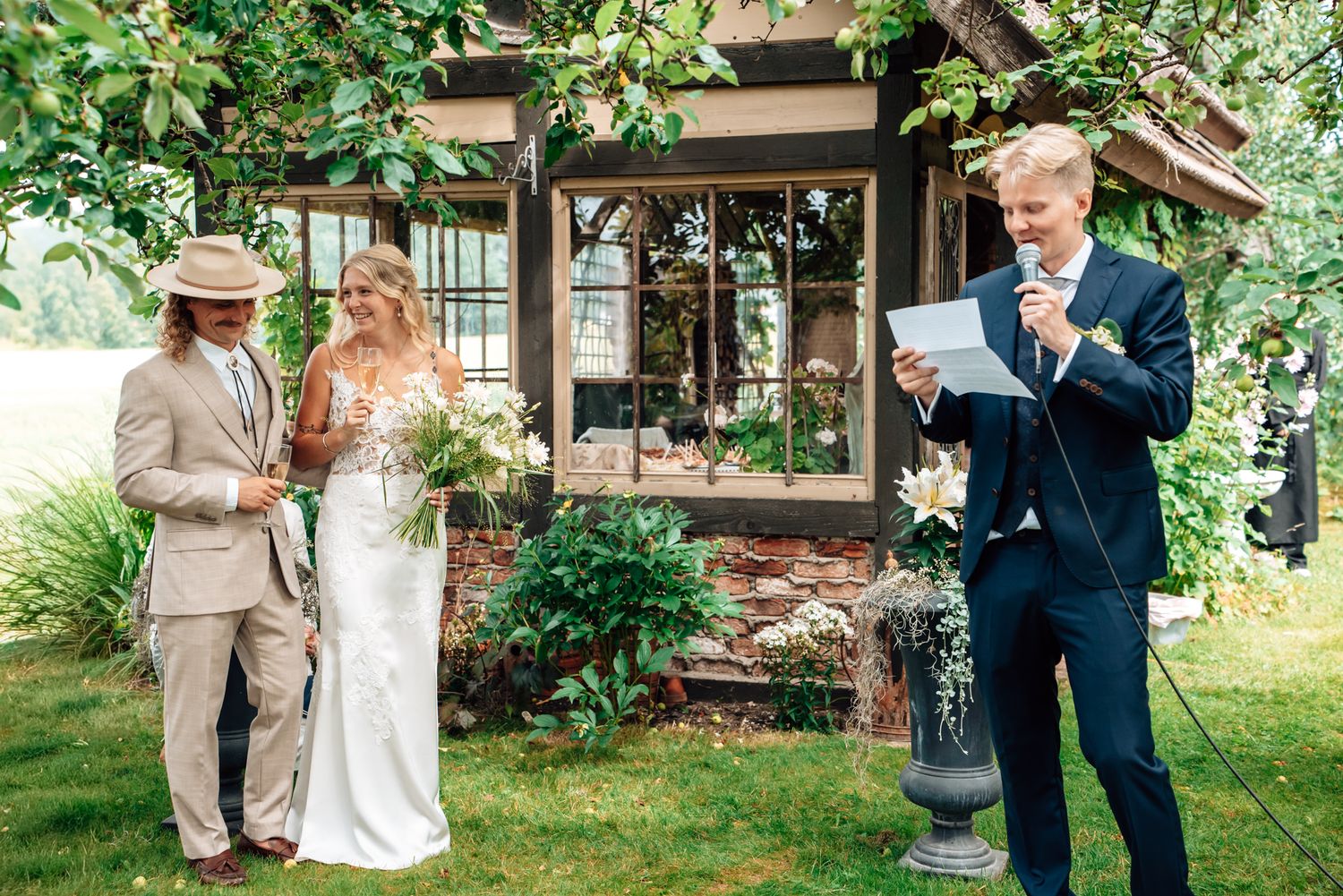 Outdoor wedding ceremony with bride, groom, and officiant reading vows near a rustic building surrounded by lush greenery.