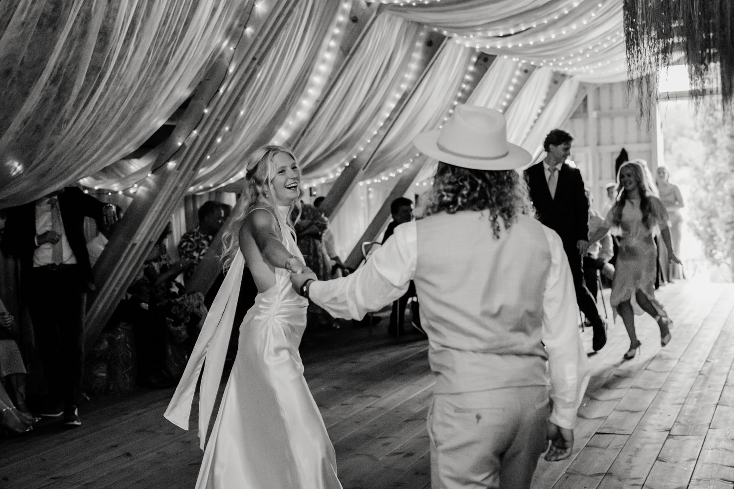 Black and white photo of bride and groom sharing first dance at elegant wedding reception with draped fabric and string lights.