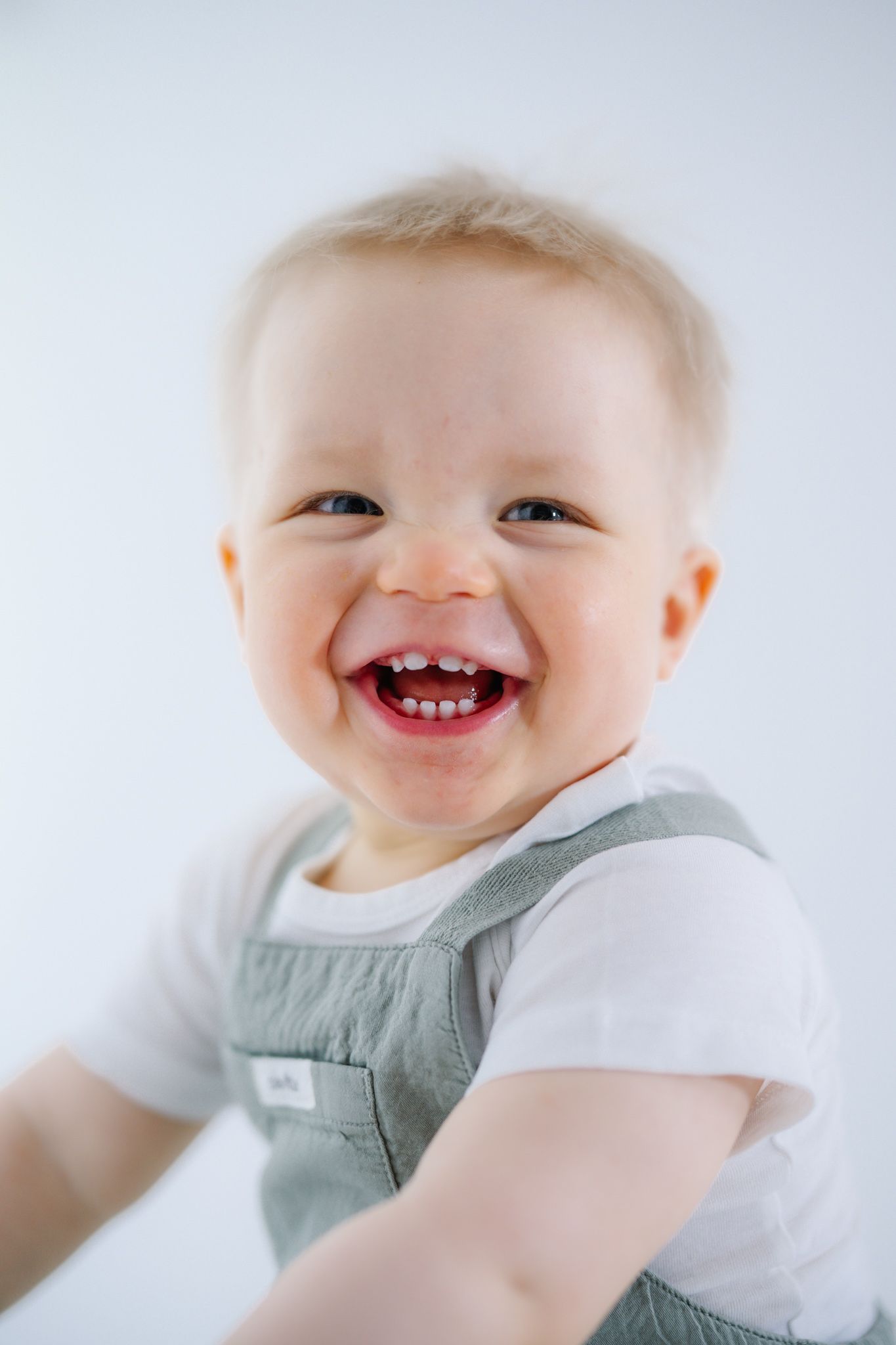 A joyful baby with a big smile wearing a white shirt and gray overalls against a light background.