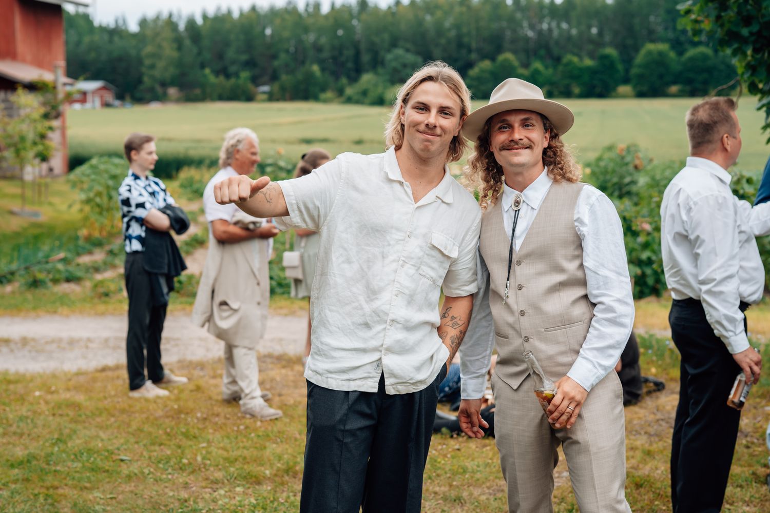 Two smiling men pose outdoors at a summer wedding celebration in a lush green countryside setting.