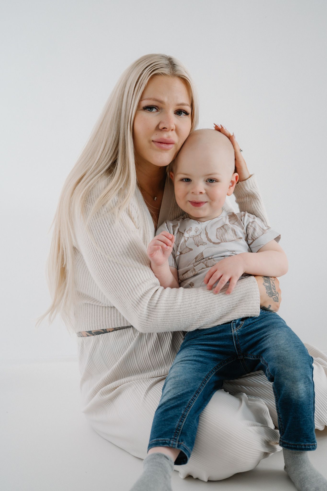 A blonde woman in a white cardigan sits on a pouf, lovingly holding a smiling baby in jeans against a white background.