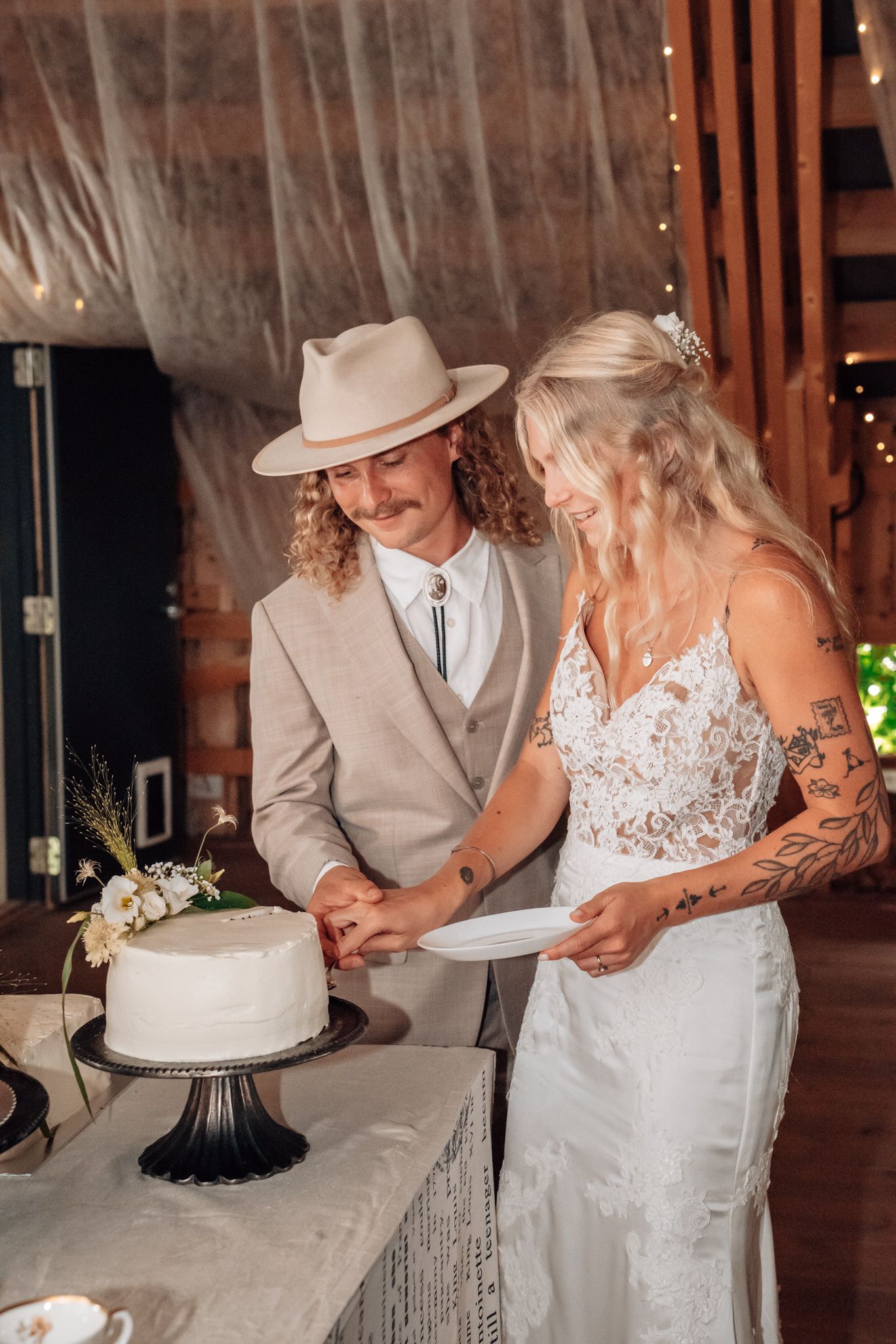 Bride and groom cutting white wedding cake together in rustic barn venue, groom wearing tan suit and wide-brim hat.