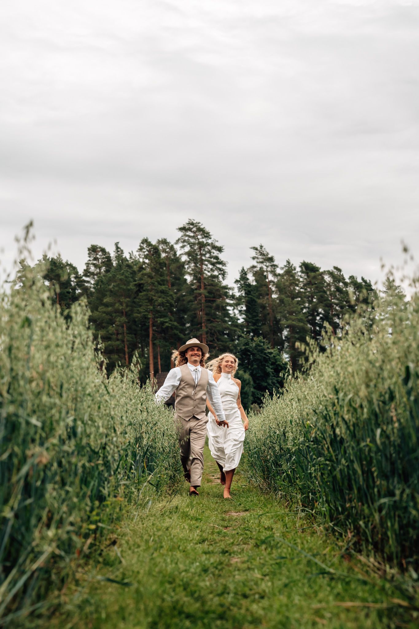 Happy couple running through a lush green field path with pine trees in background on a cloudy day.