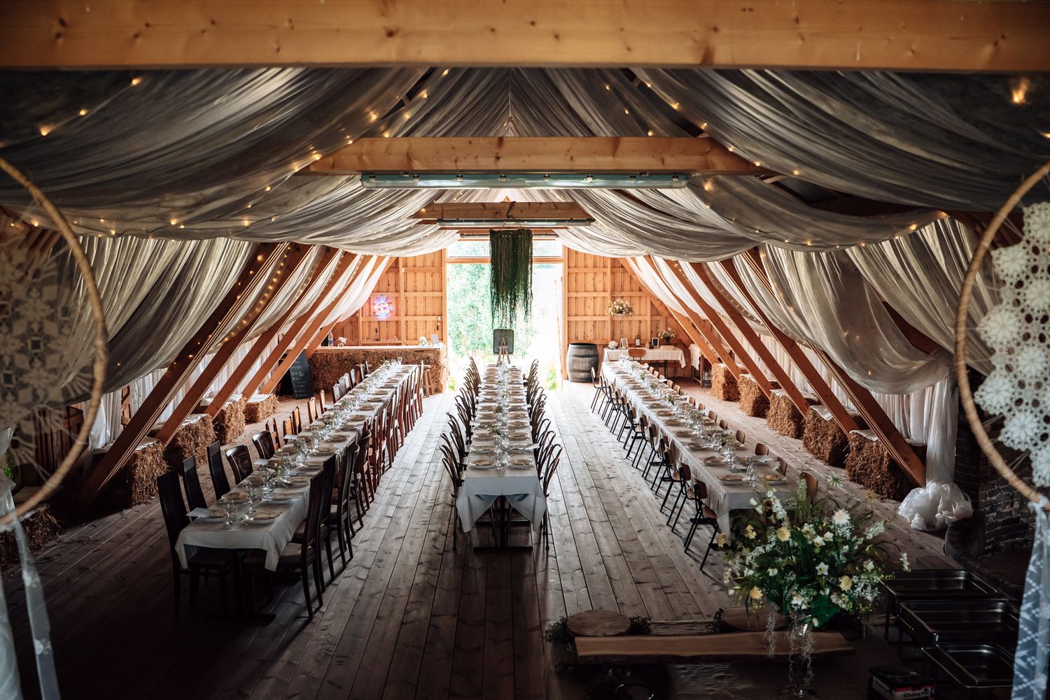 Rustic barn wedding reception with long decorated tables, fairy lights, and elegant draped fabric ceiling.