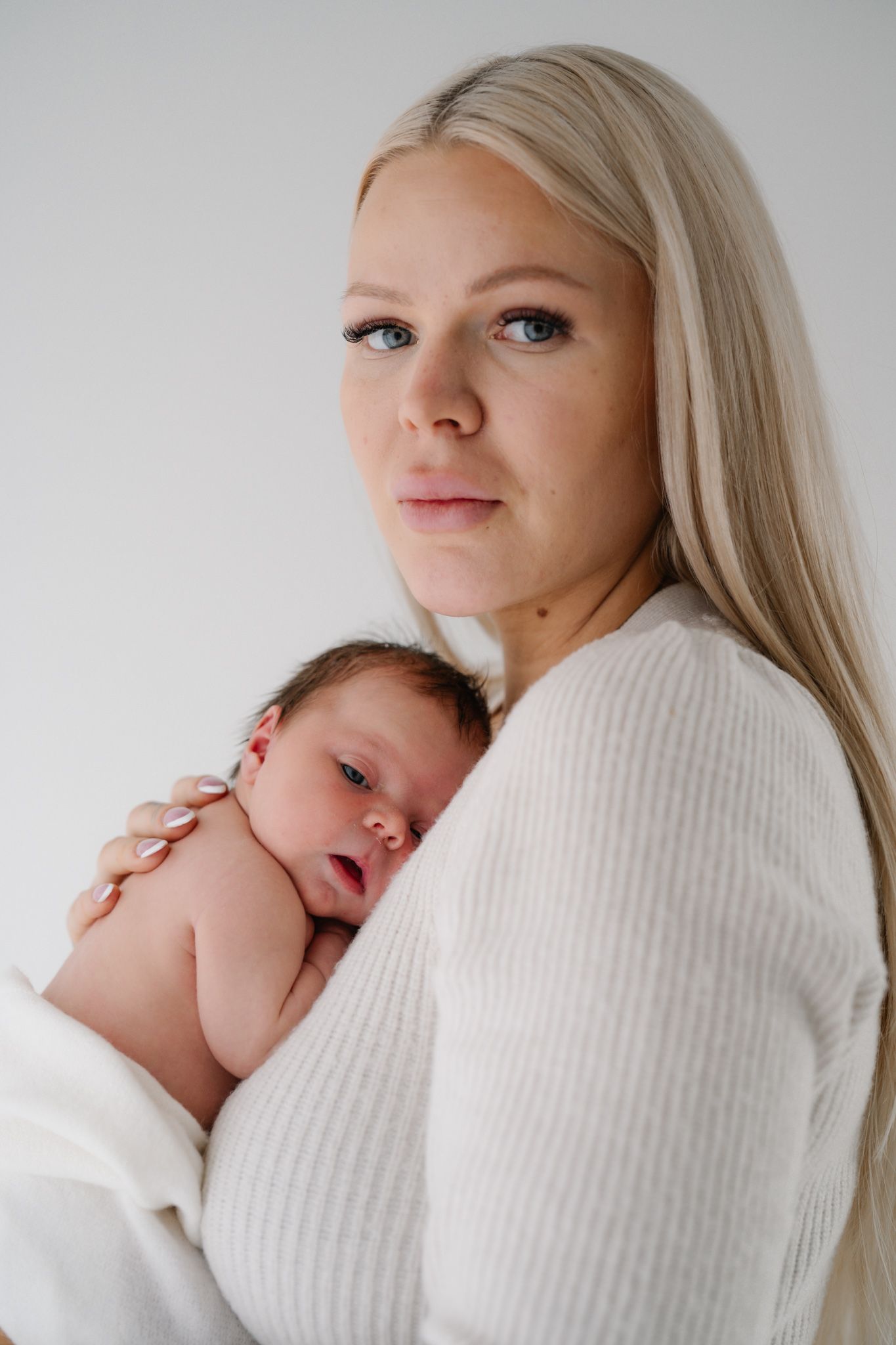 A blonde woman with blue eyes in a white sweater tenderly holds a sleeping newborn baby against her chest.