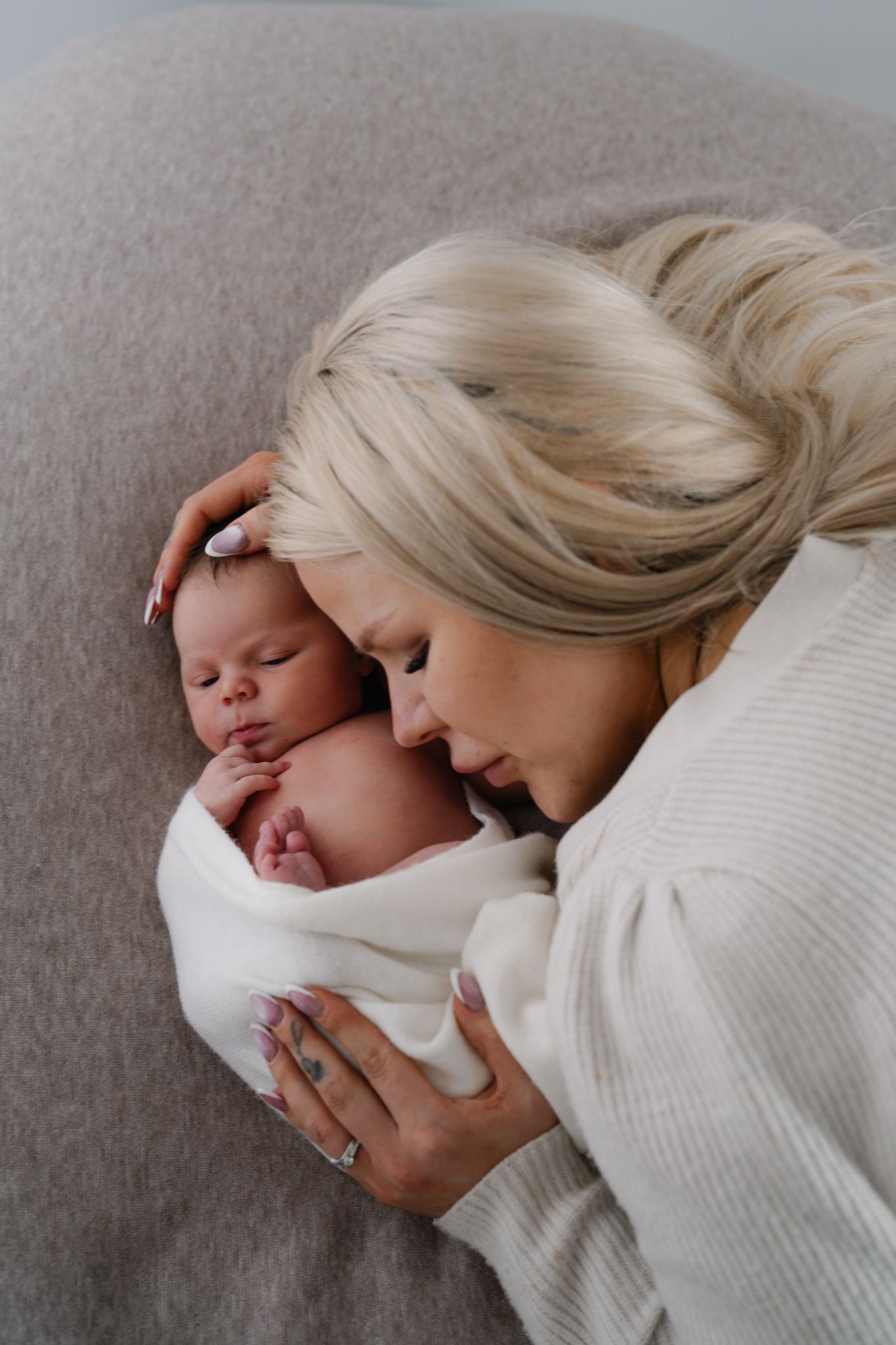 Blonde mother tenderly nuzzling her sleeping newborn baby wrapped in a white swaddle blanket.
