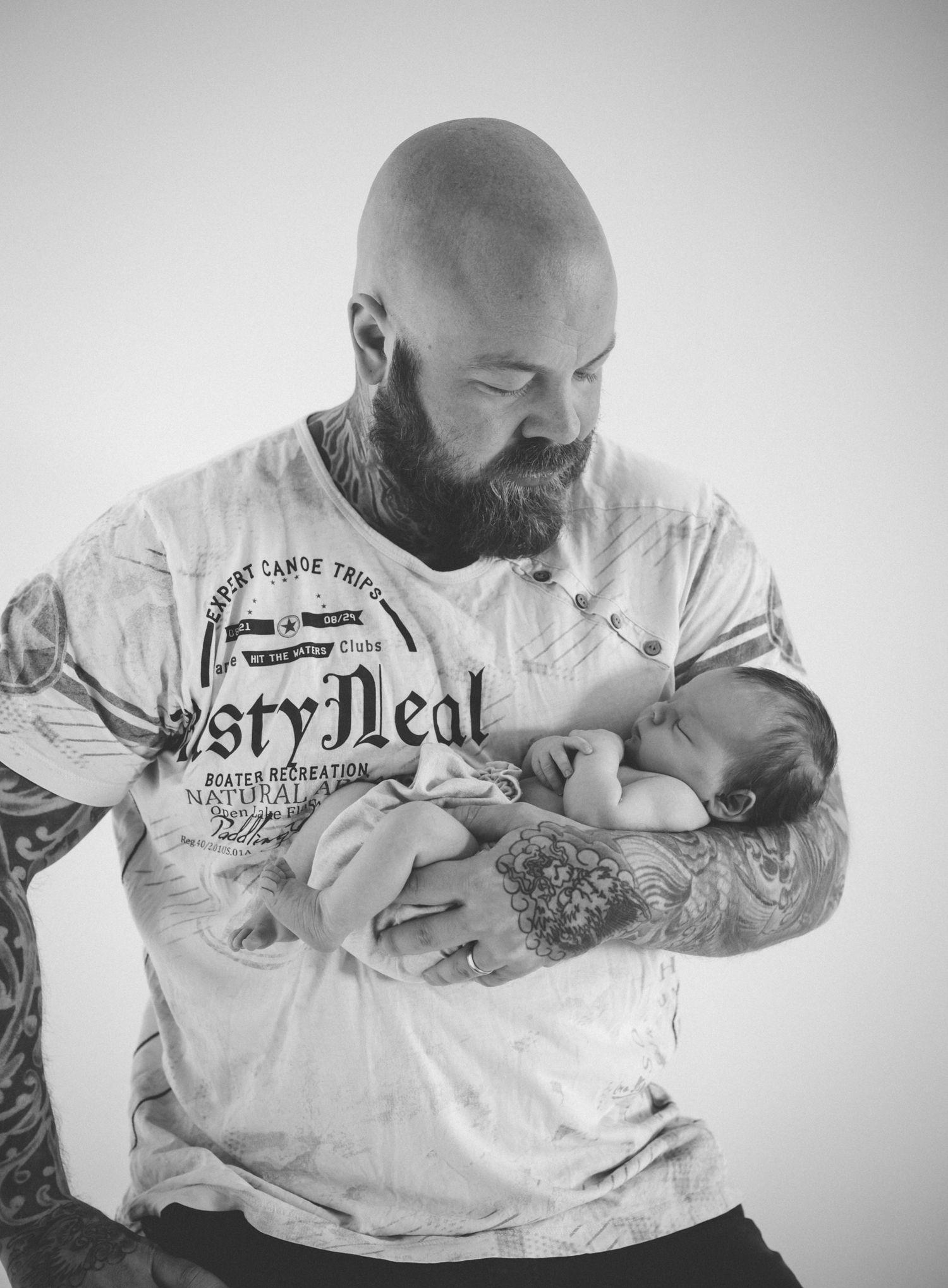Bald tattooed man with beard cradling a sleeping newborn baby in his arms, black and white portrait photo.