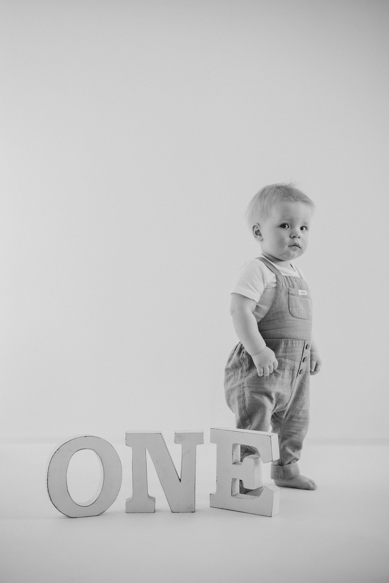 Black and white photo of a one-year-old baby standing next to wooden ONE letters for a first birthday portrait.