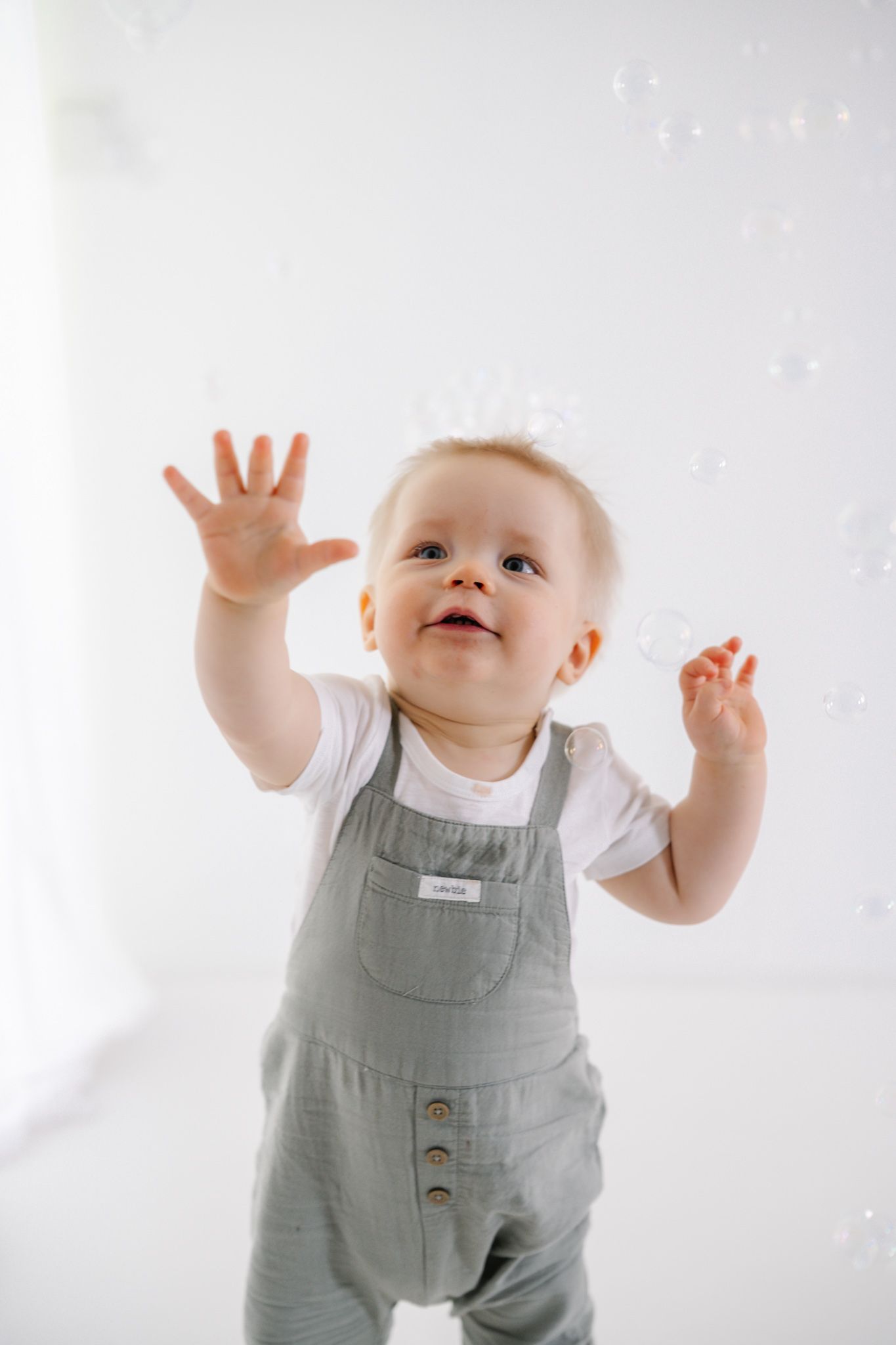 A curious baby in gray overalls reaches up toward floating soap bubbles against a white background.