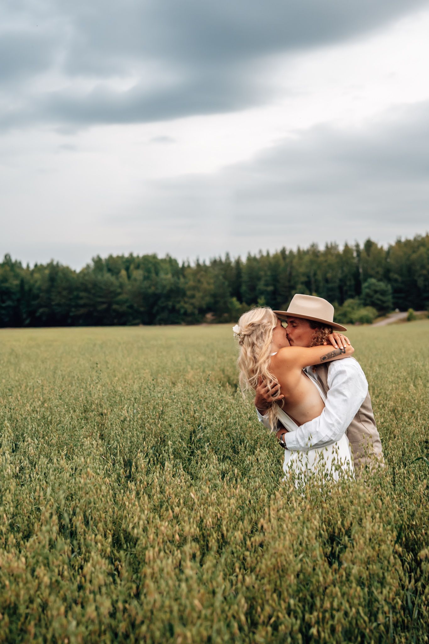 Couple embracing and kissing in a lush green field with a forest backdrop under a cloudy sky.
