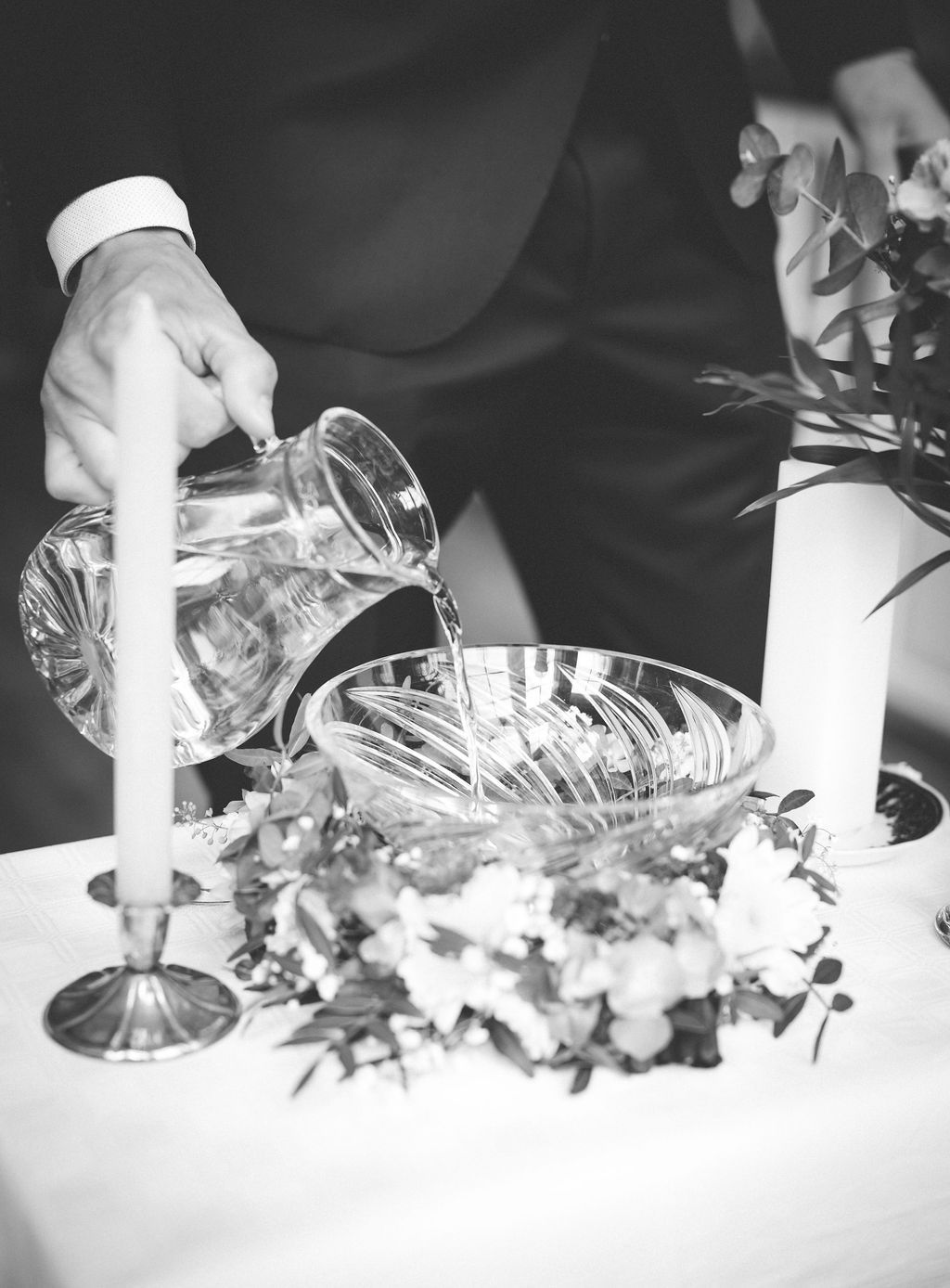 Black and white photo of a person pouring water into a crystal bowl during a wedding ceremony with candles and flowers.
