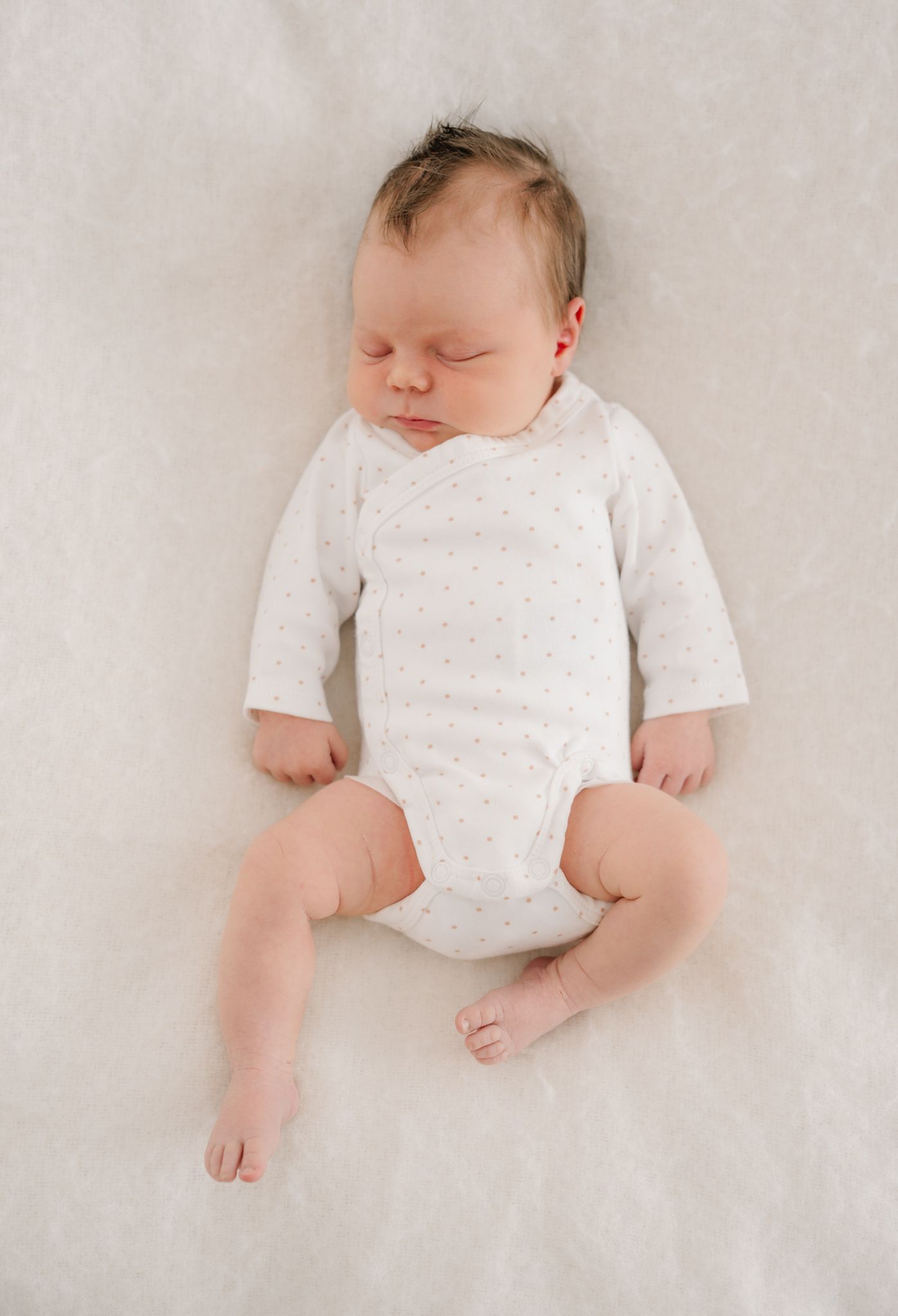 Peaceful newborn baby sleeping on white fluffy blanket wearing a white long-sleeve dotted onesie.