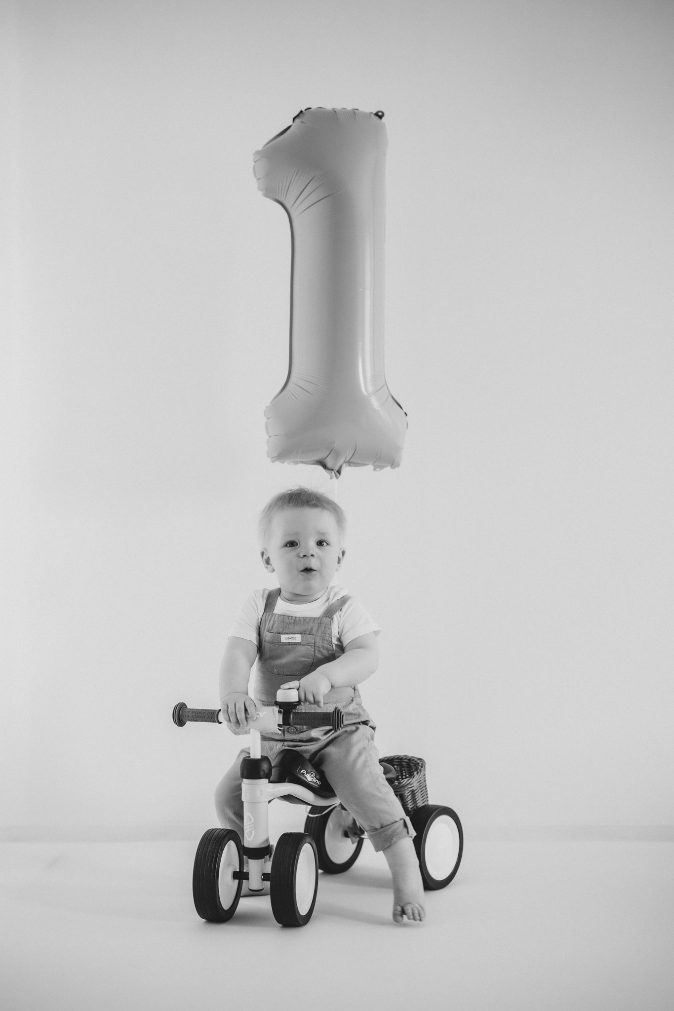 Black and white photo of a baby on a tricycle celebrating their first birthday with a number one balloon.
