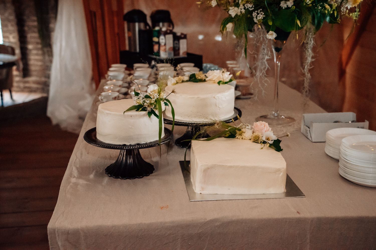 Elegant wedding dessert table with white frosted round and square cakes decorated with fresh flowers at a reception.