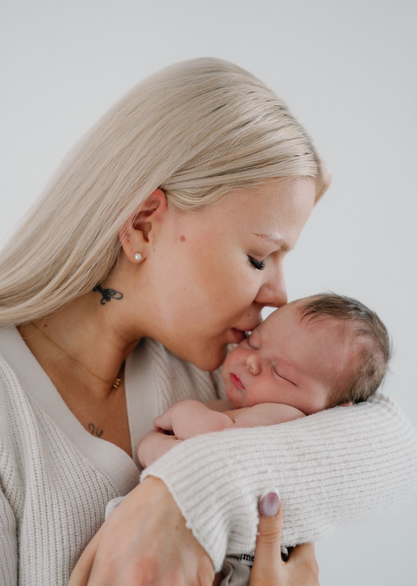 A mother with blonde hair gently kisses her sleeping newborn baby wrapped in a white blanket.