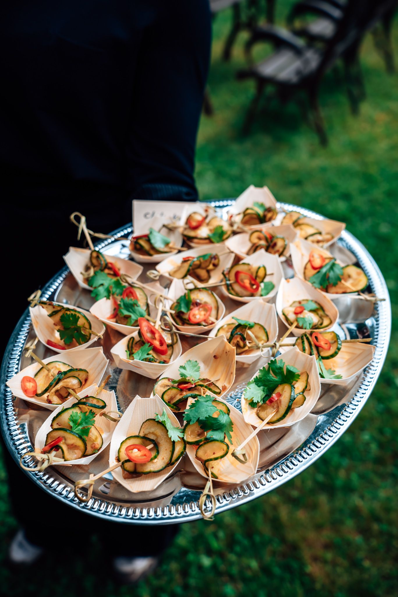A waiter holds a silver tray of elegant appetizers in paper cups, topped with zucchini, tomatoes, and fresh cilantro.