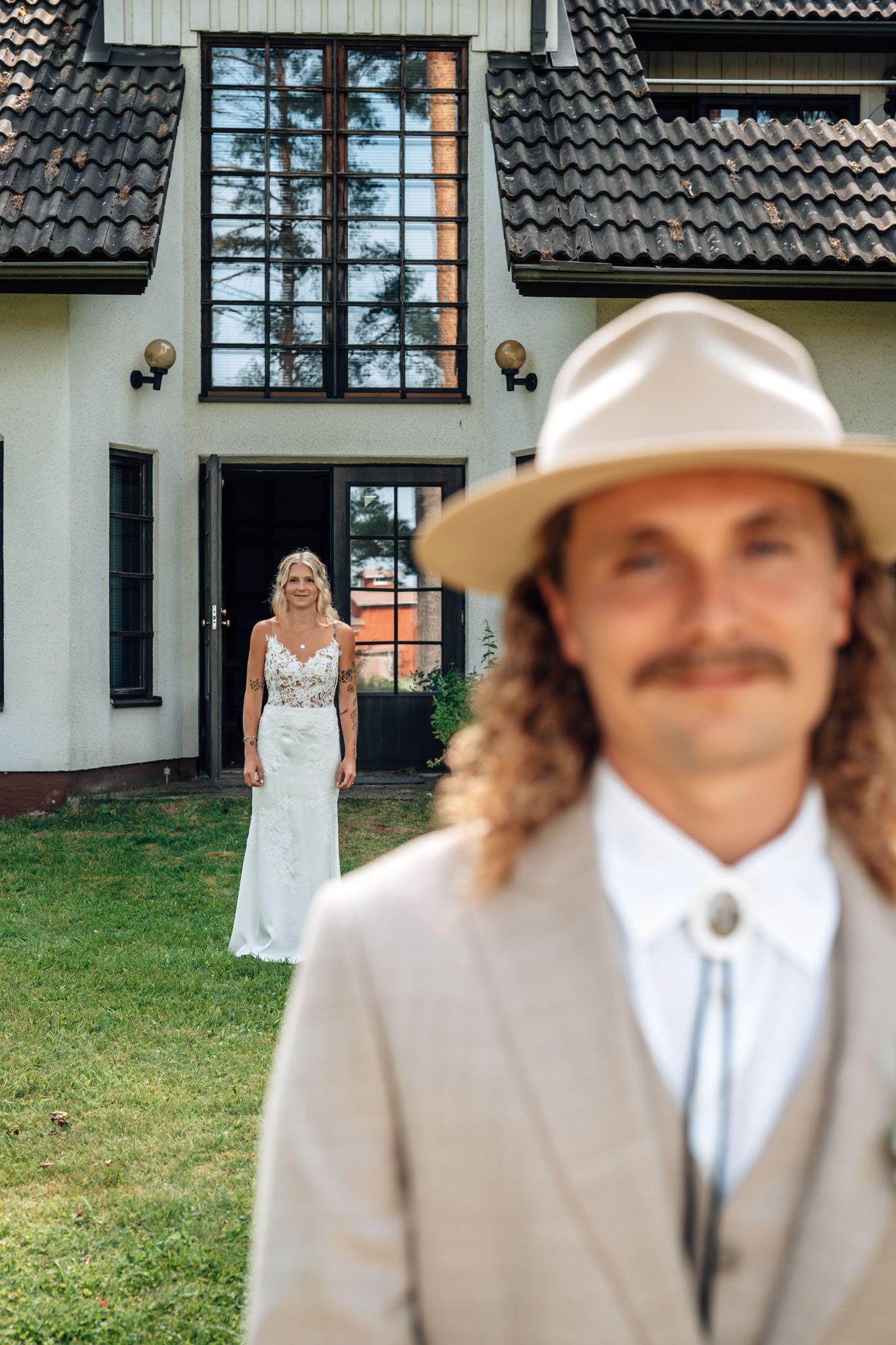 A groom in a tan suit and cowboy hat stands in foreground as his bride in a white lace gown poses by a house.