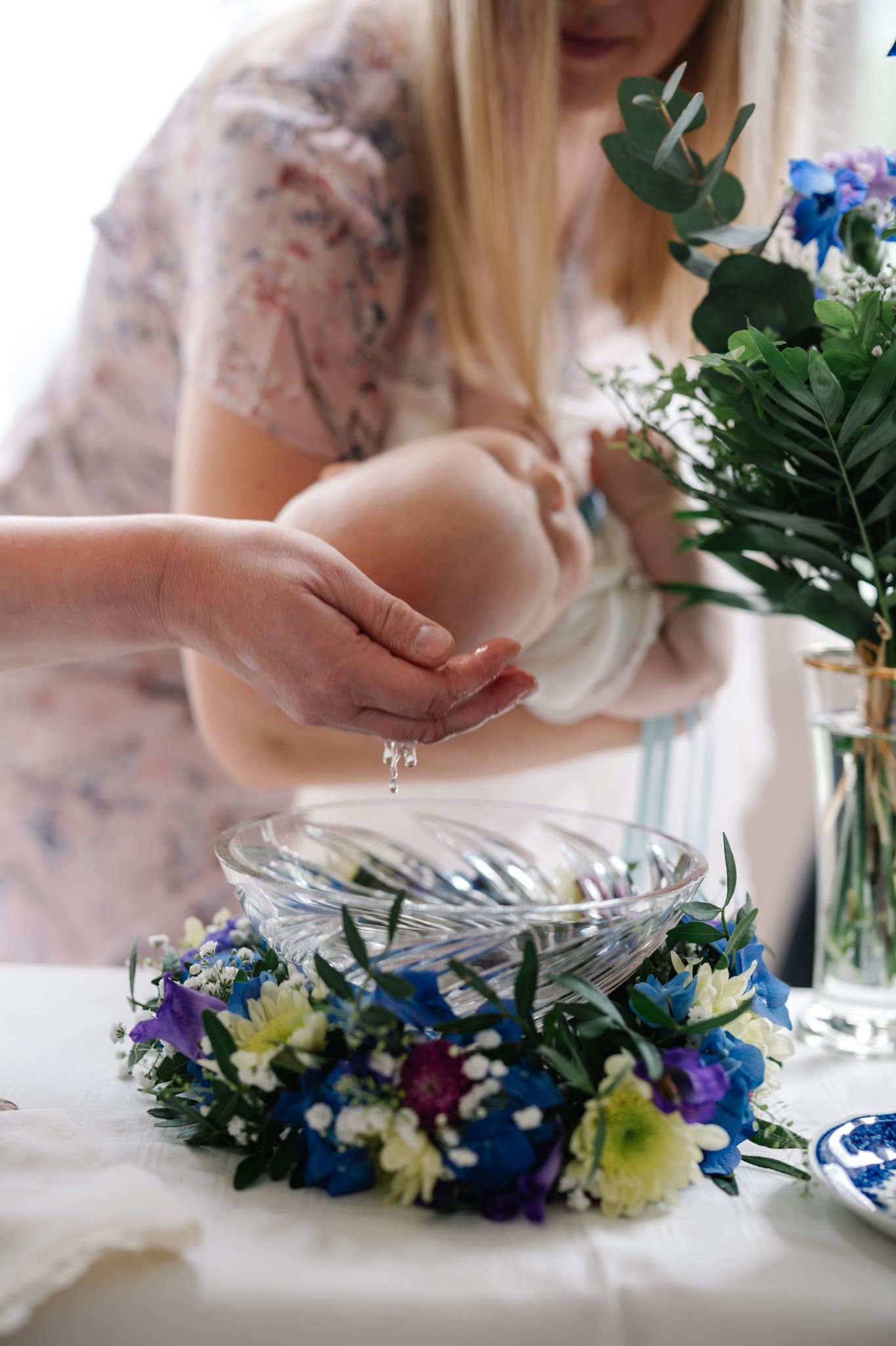 A baby baptism ceremony with water droplets falling into a glass bowl surrounded by a floral wreath on a white table.