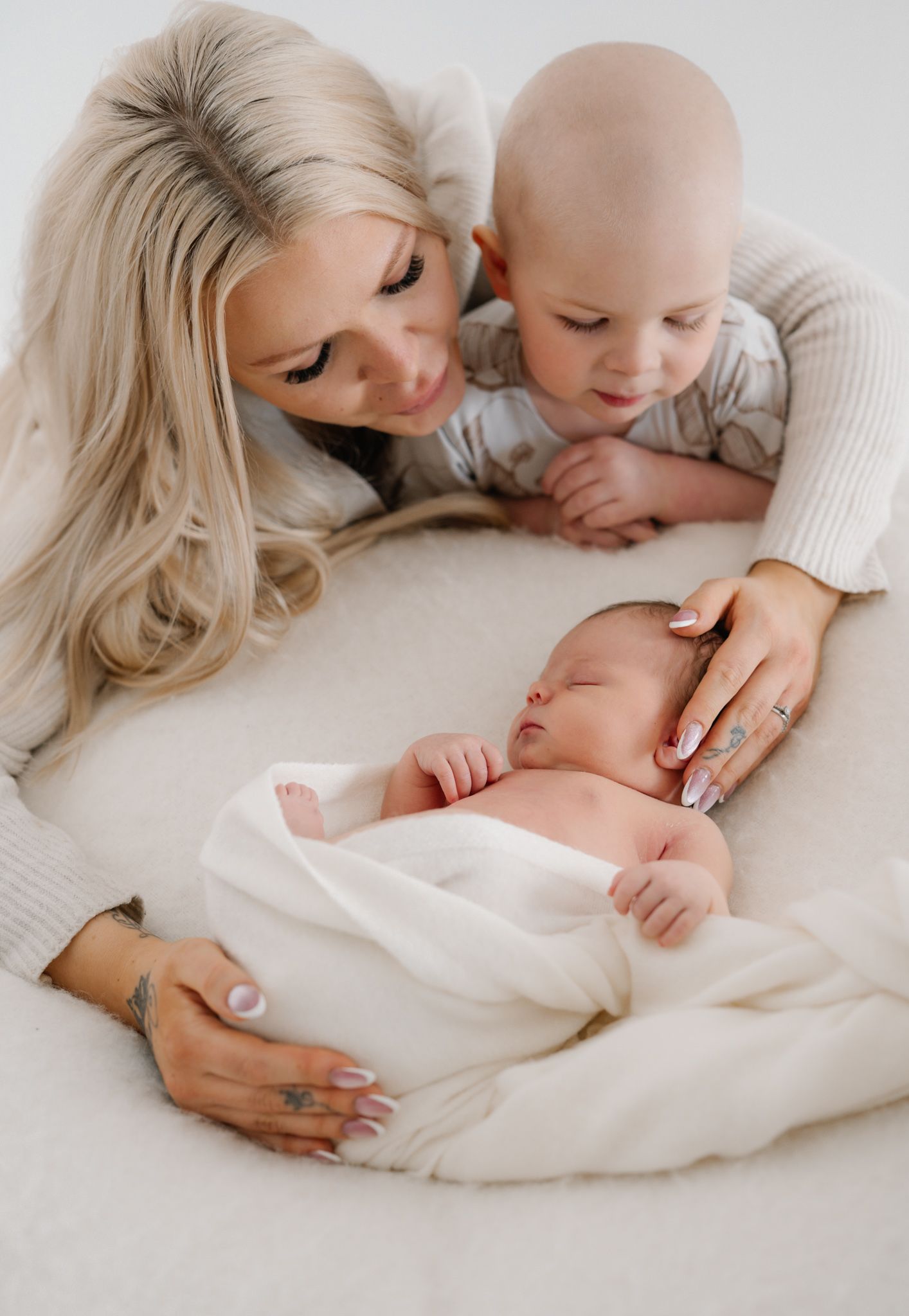 A mother with blonde hair gazes lovingly at her newborn baby wrapped in white, with a toddler leaning in beside her.