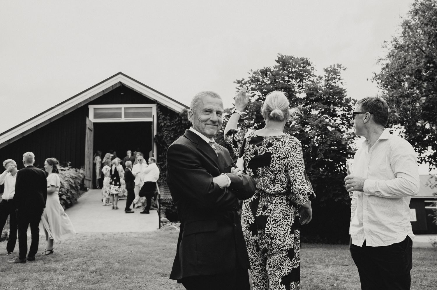 Black and white photo of elegantly dressed guests socializing outdoors at a wedding reception near a rustic barn.