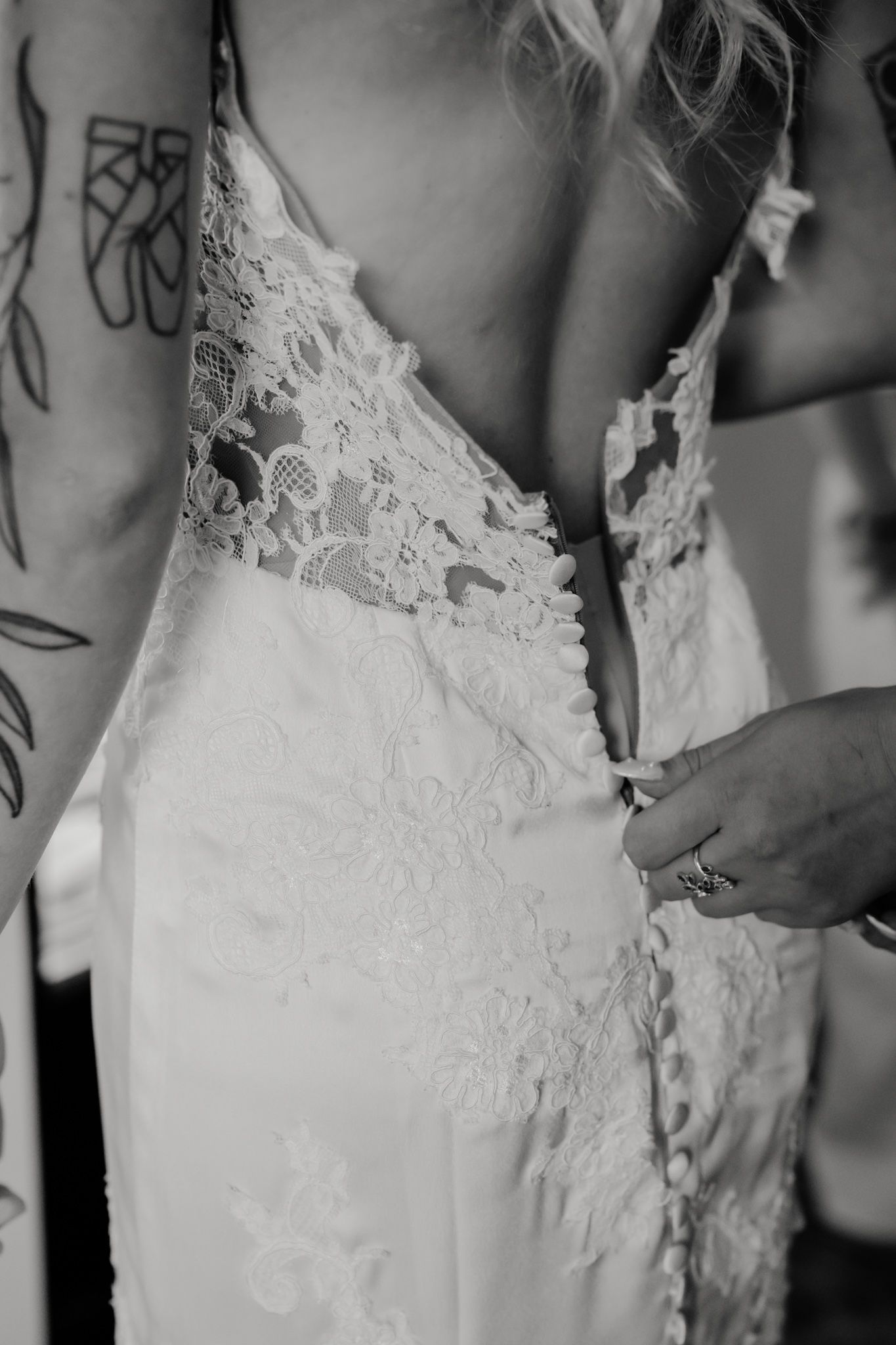 Black and white photo of a bride in a lace backless wedding gown being buttoned up by a helper.