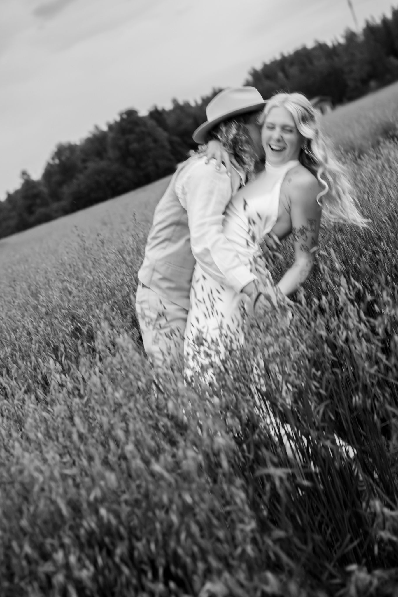 Black and white photo of a laughing couple walking through a field, woman in floral dress, man in hat.