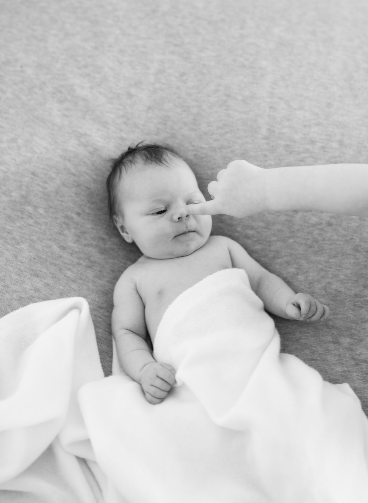 Black and white photo of a newborn baby wrapped in white blanket with parent's hand gently touching nose.