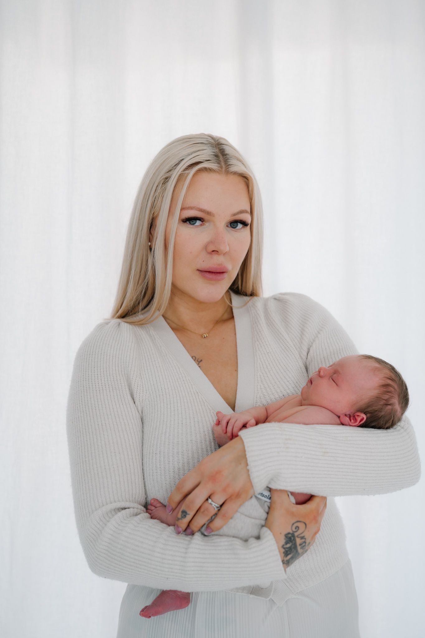 Blonde woman in cream ribbed wrap dress tenderly holding sleeping newborn baby against white curtain backdrop.