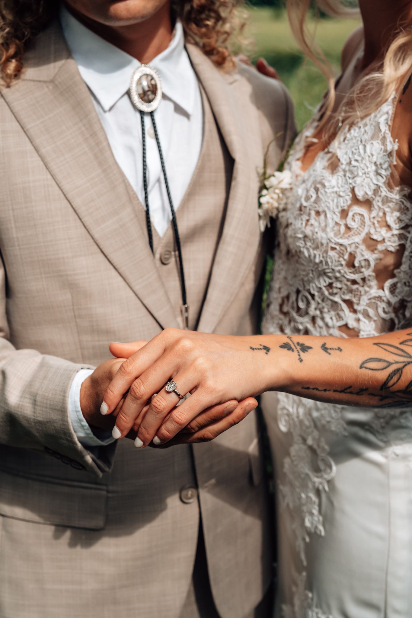 Bride and groom holding hands at wedding, bride wearing diamond ring and floral tattoo, groom in beige suit with bolo tie.