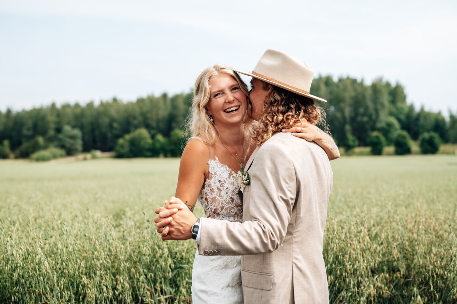 A joyful bride and groom dancing in a lush green field, the bride laughing as the groom wears a tan hat.