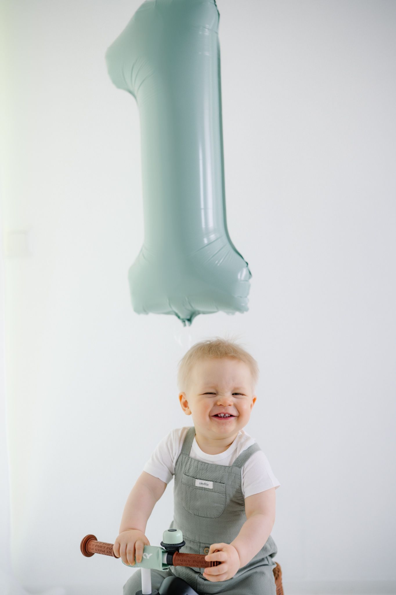 Smiling baby boy on a balance bike celebrating his 1st birthday with a light blue number one balloon.