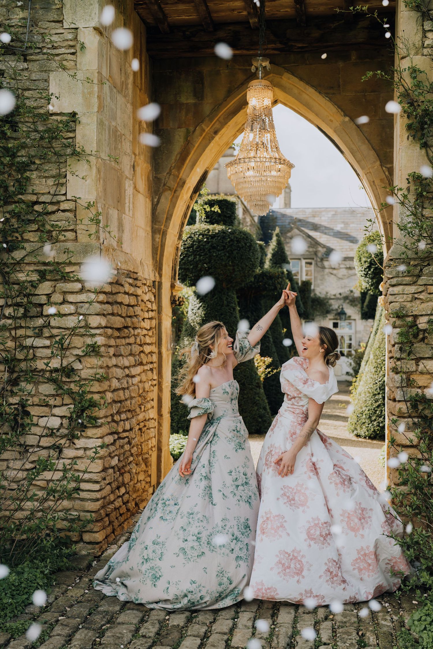 Two brides in floral gowns celebrate under a stone arch with chandelier, confetti falling at elegant castle venue.