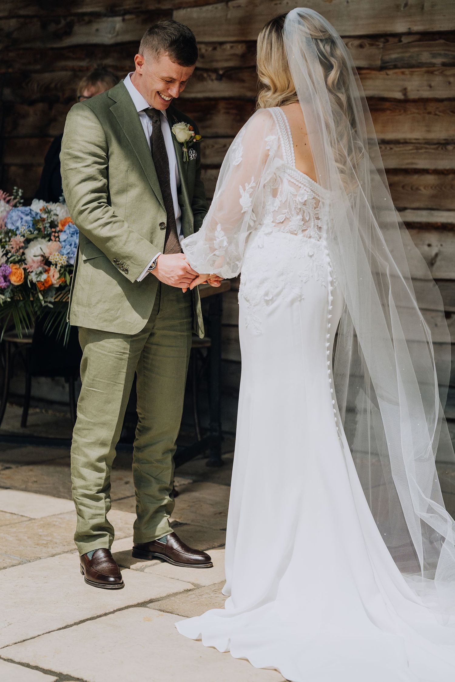 Groom in olive green suit holding bride's hands during outdoor barn wedding ceremony.