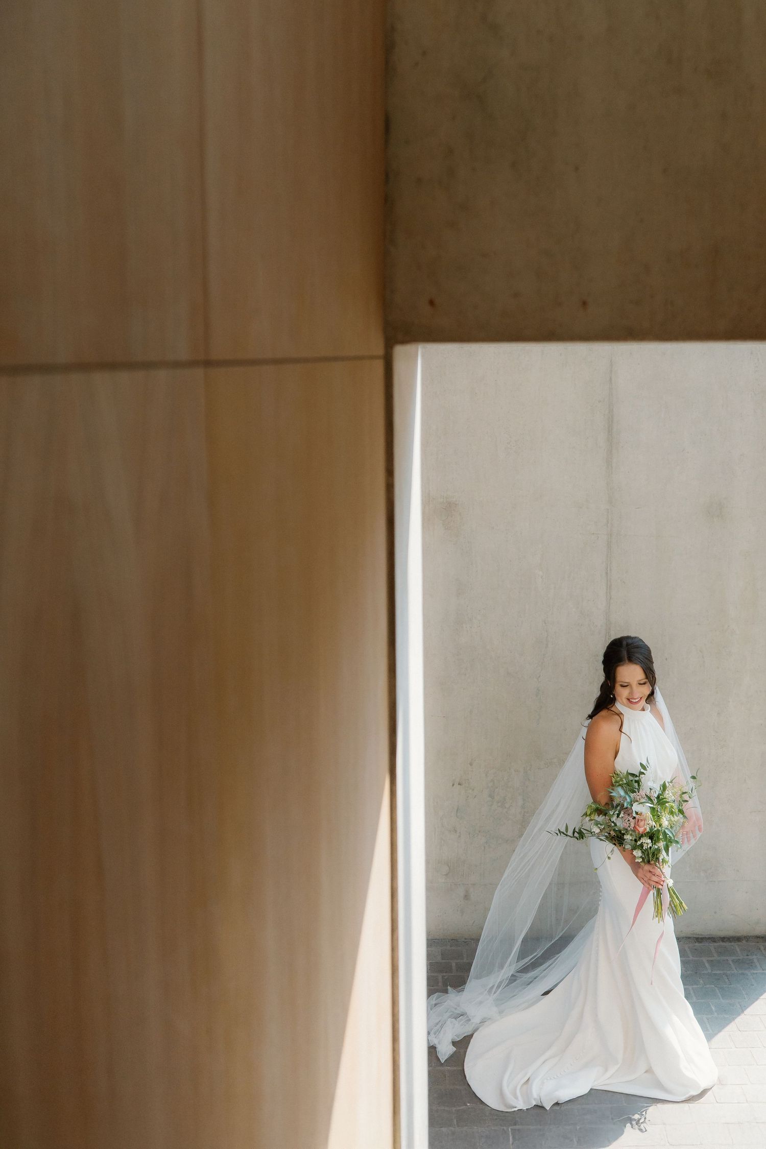 Bride in flowing white gown and veil holding bouquet framed by modern architectural concrete columns.