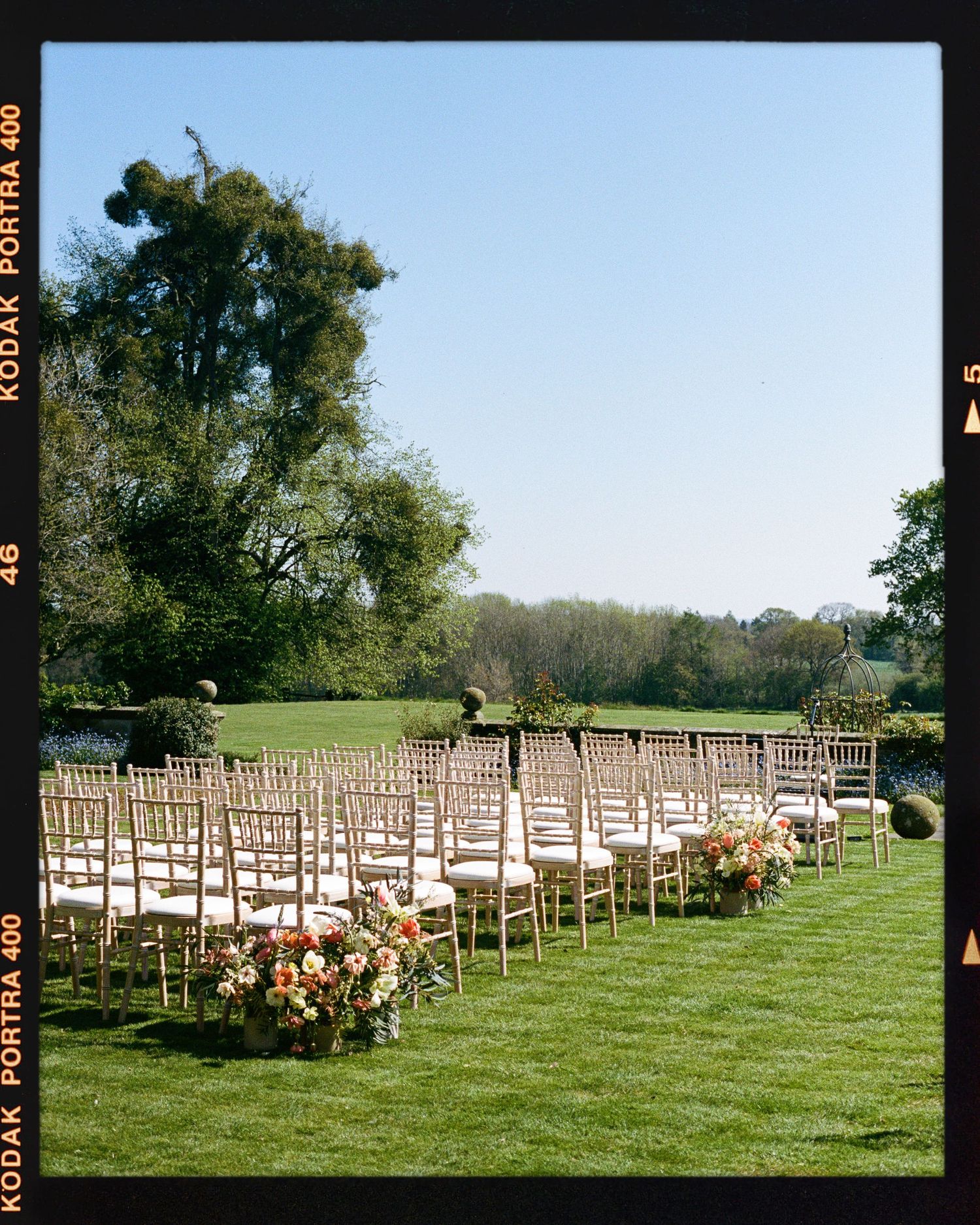 Outdoor wedding ceremony setup with gold chiavari chairs arranged on lush green lawn, adorned with colorful floral arrangements.