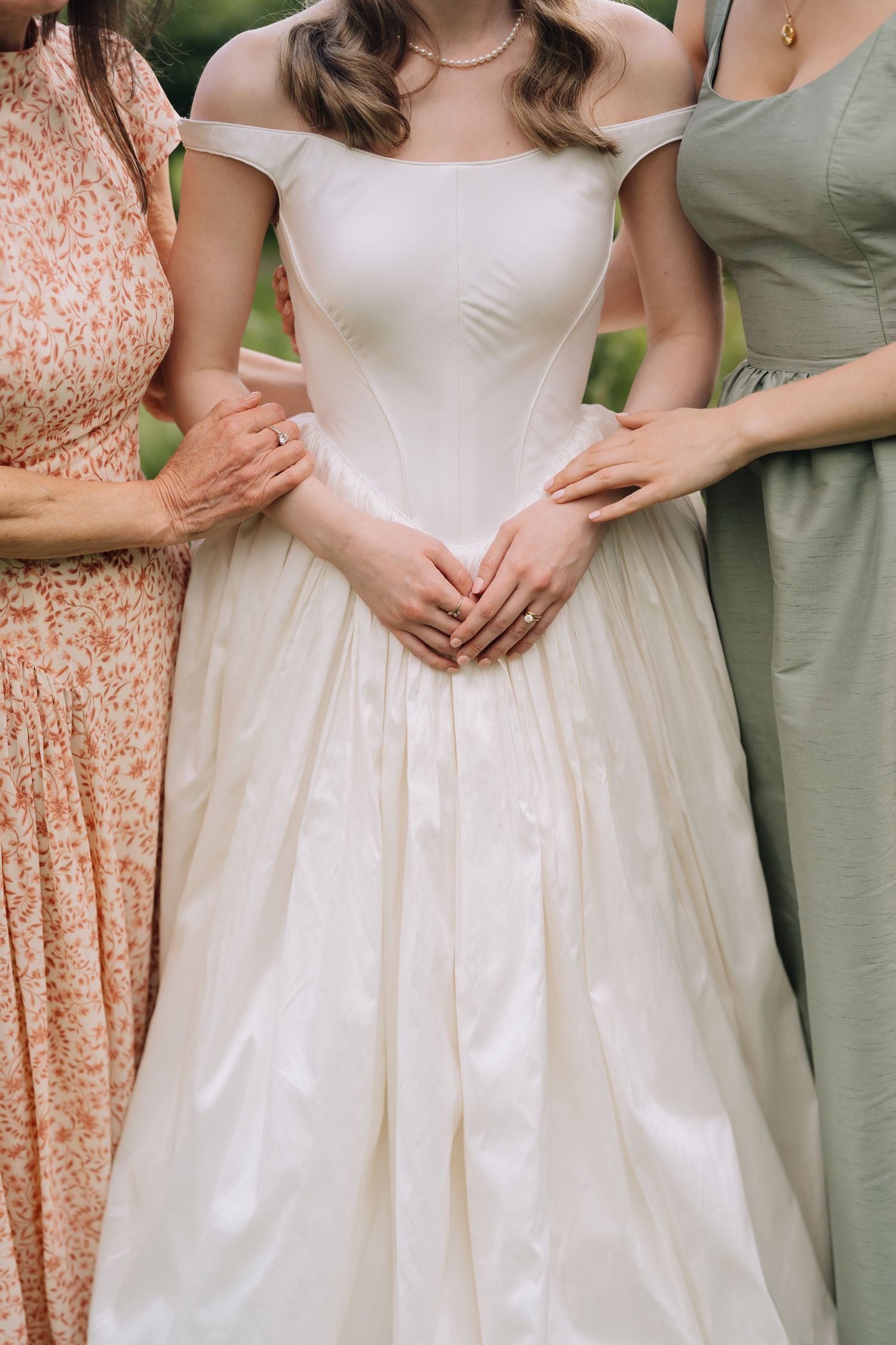 Bride in elegant off-shoulder white ball gown flanked by two women in floral and sage dresses outdoors.