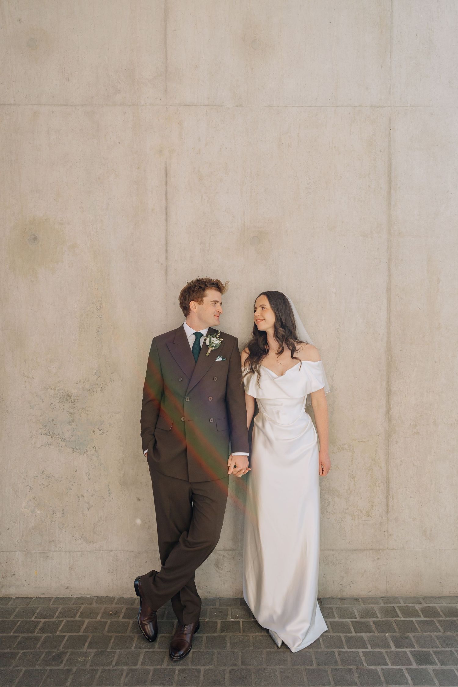 A bride and groom hold hands smiling at each other, posing against a concrete wall in elegant wedding attire.